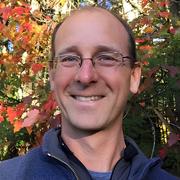 Professional headshot of a man with glasses smiling in front of vibrant fall foliage